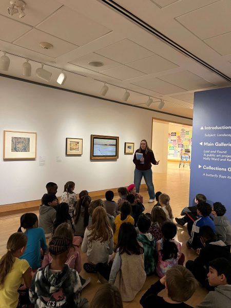 School group sitting on gallery floor looking at paintings on the walls while a guide speaks to them